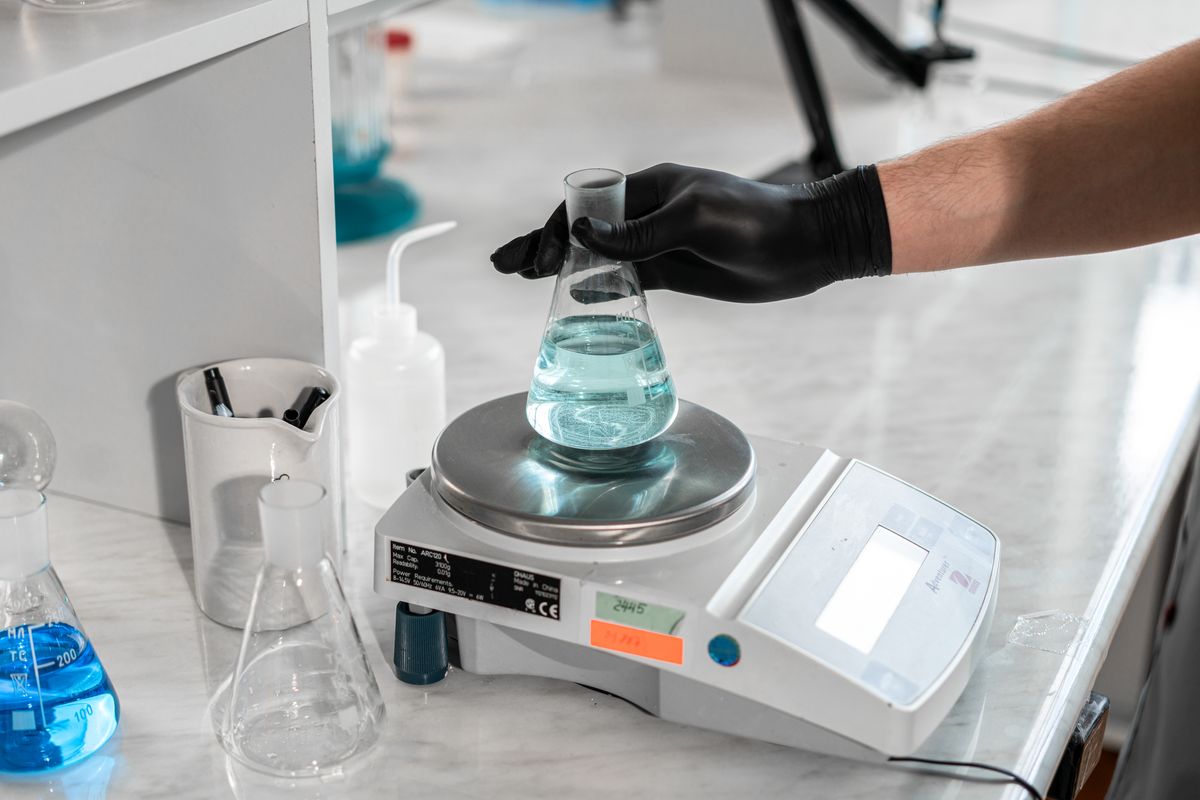 A scientist handling a liquid-filled flask on a laboratory scale for precise measurement.