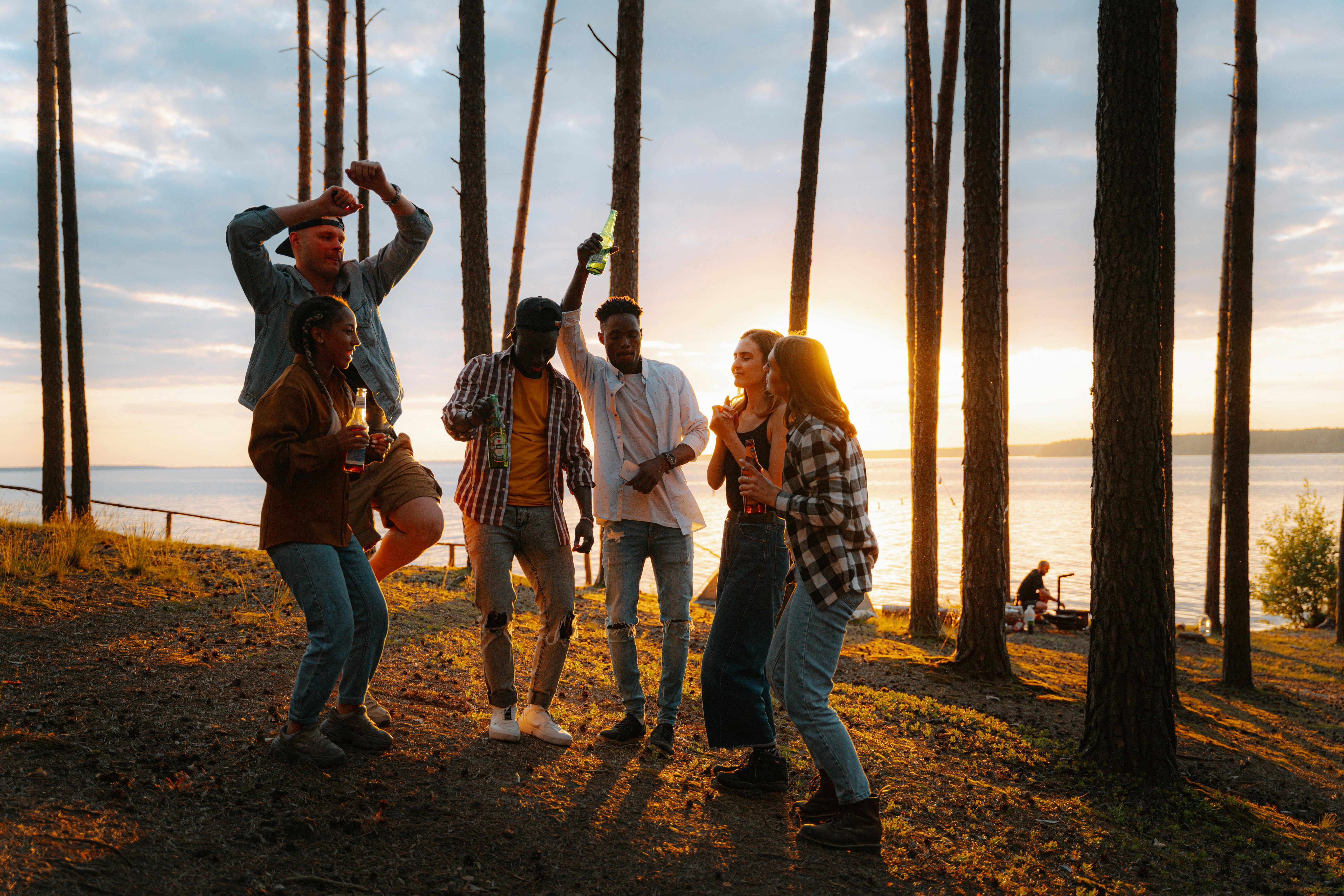 Cheerful group of young friends dancing outdoors at sunset near a lake, enjoying nature.