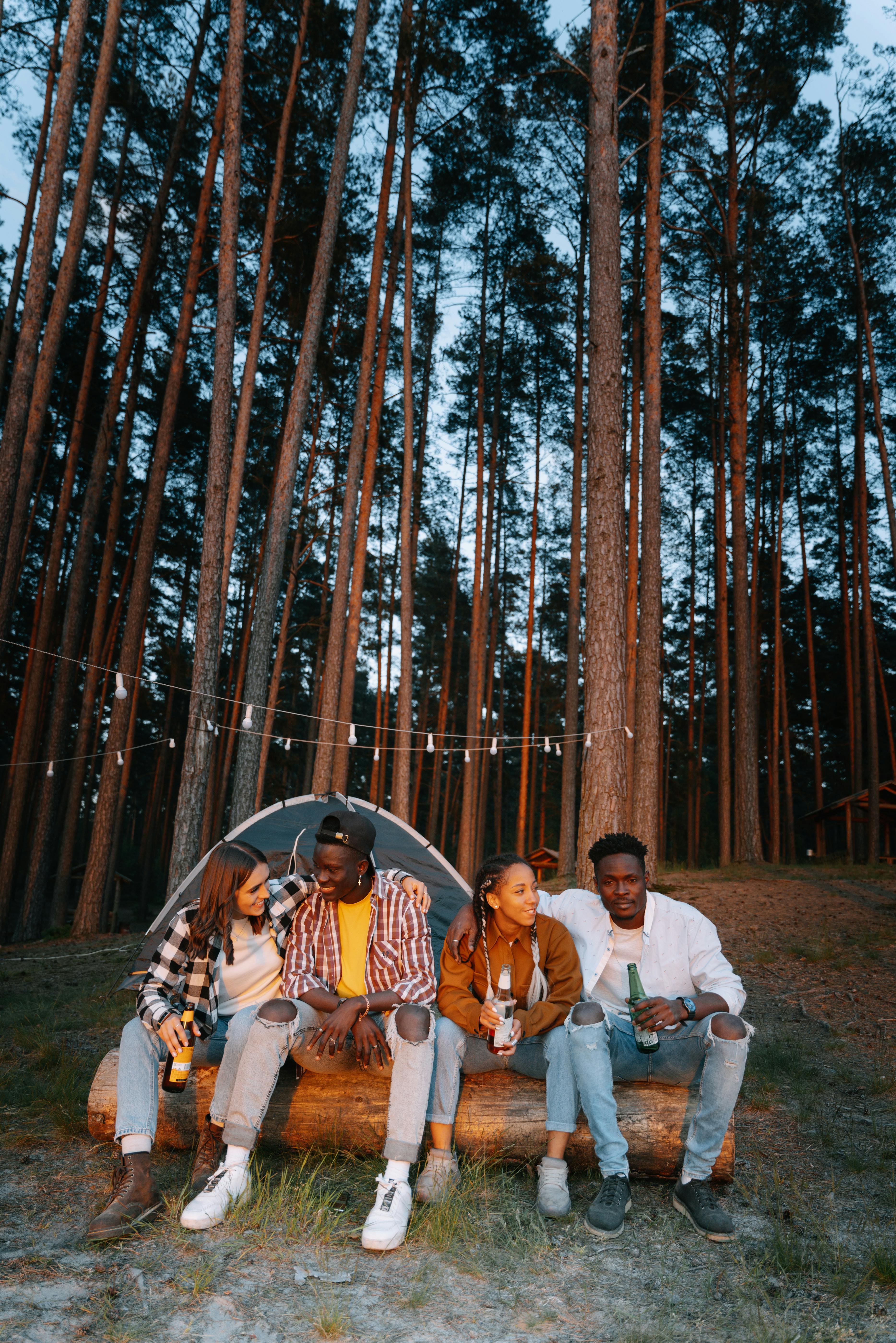 Four friends relax on a log with drinks, surrounded by nature and tall trees during a camping trip.