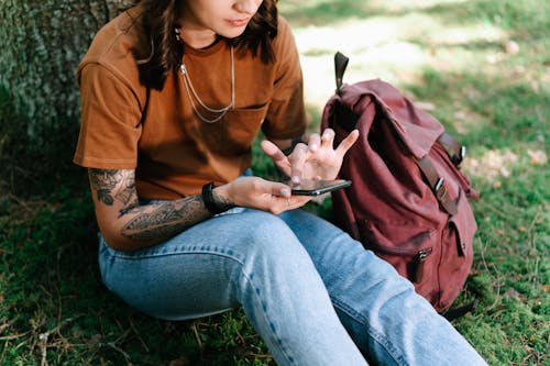 A Tattooed Woman Using Her Smartphone while Sitting on the Grass