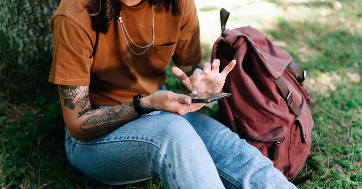 Mulher tatuada sentada sob uma árvore usando seu smartphone com uma mochila ao lado.