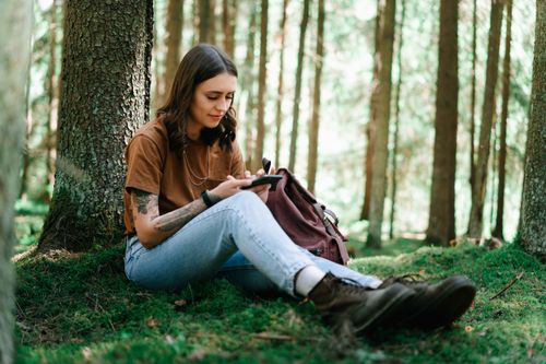 Free Young woman sitting in a forest using a smartphone, surrounded by nature. Relaxation and adventure. Stock Photo