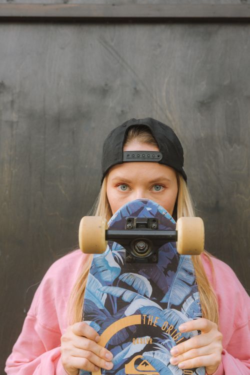 Free Portrait of a young woman with a skateboard, wearing a cap, standing outdoors against a dark wall. Stock Photo