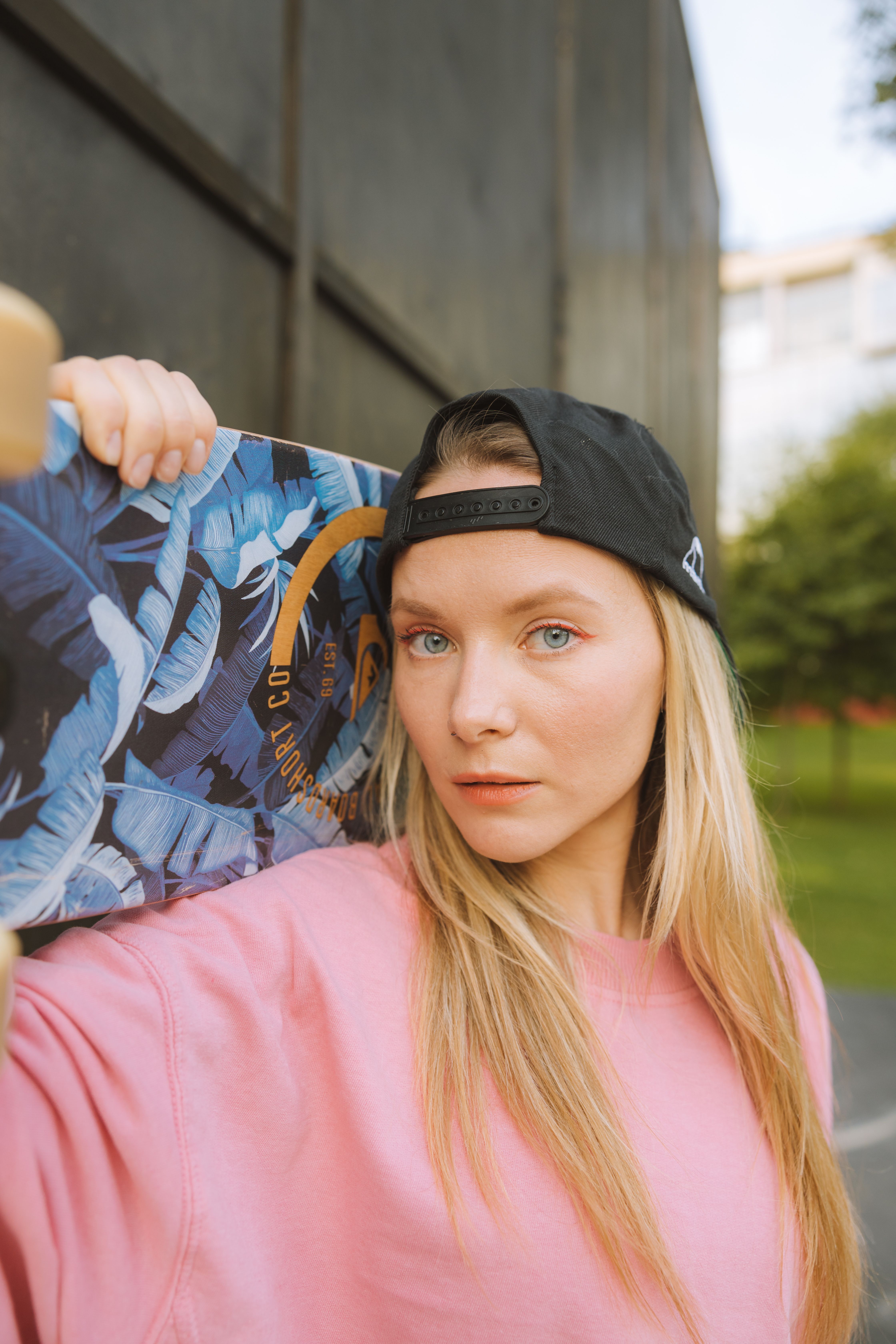 Blonde woman with skateboard in park wearing pink shirt and black cap.