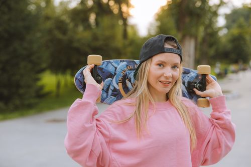 Free Blonde woman in pink pullover holds a skateboard in a park, showcasing relaxed summer vibes. Stock Photo