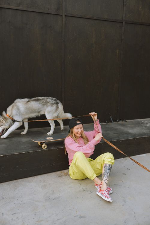 Free A cheerful young woman sitting on a sidewalk, playfully engaging with her dog on a leash beside a skateboard. Stock Photo