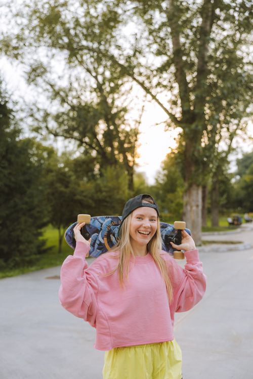Free Smiling woman in a pink sweater holding a skateboard on her shoulders outdoors. Stock Photo