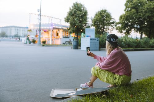 Free A teenager in casual attire taking a selfie with a skateboard beside a street food stall. Stock Photo