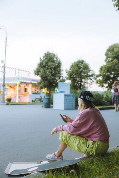 Free A young woman in casual attire sits on pavement using her smartphone next to a skateboard, enjoying the outdoors. Stock Photo