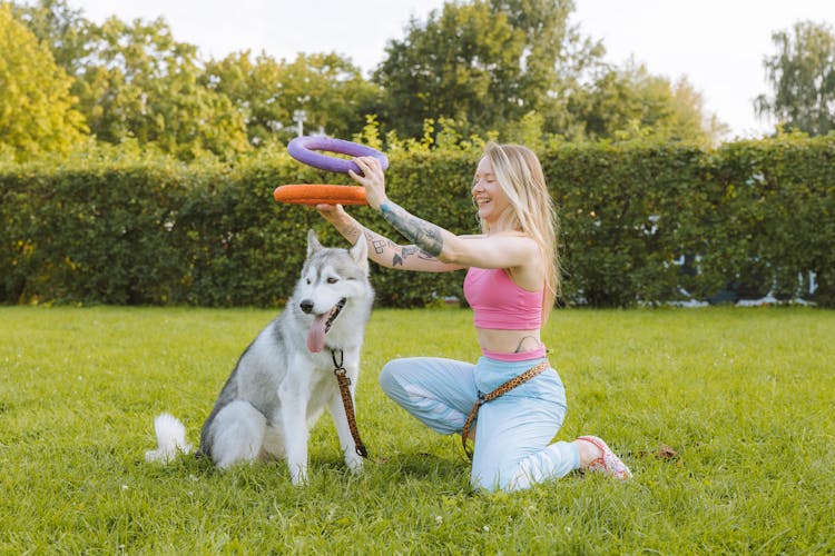 Smiling Blonde Woman With Tattoos Playing With Dog In Garden