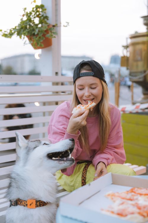 Free A young woman sharing a moment with her dog while enjoying pizza outdoors on a bench. Stock Photo