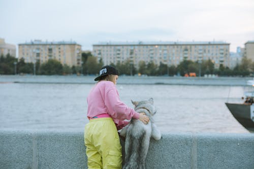 Person in Pink Shirt and Black Cap Holding a Dog Near a River