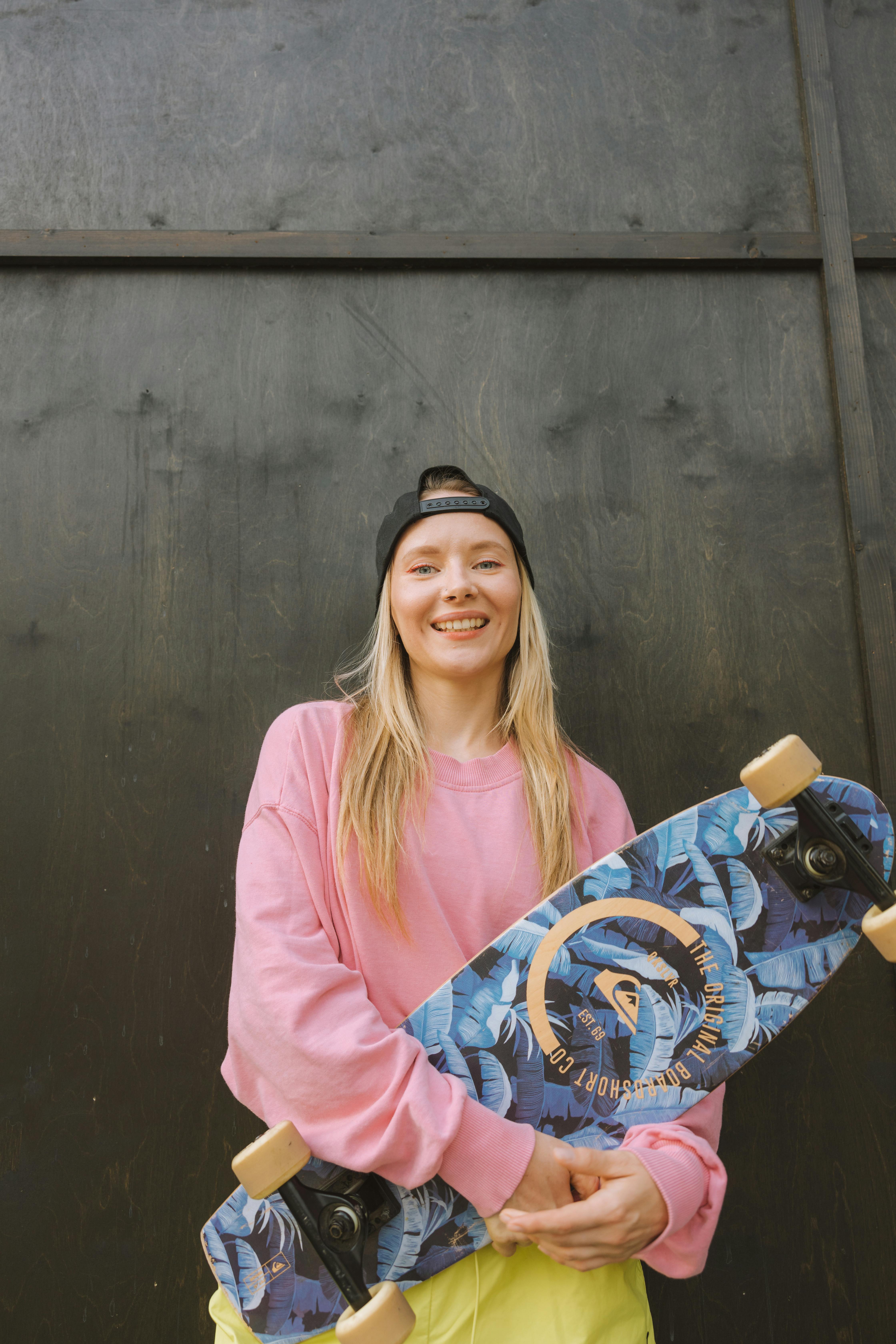 Young woman in pink sweater holding a longboard with a floral pattern, smiling at the camera.