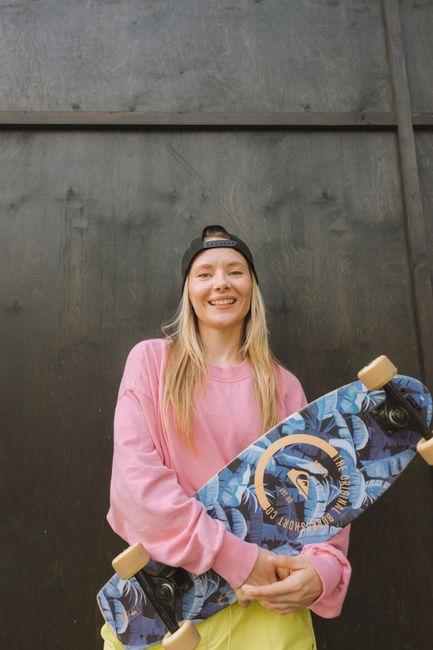 Young woman in pink sweater holding a longboard with a floral pattern, smiling at the camera.