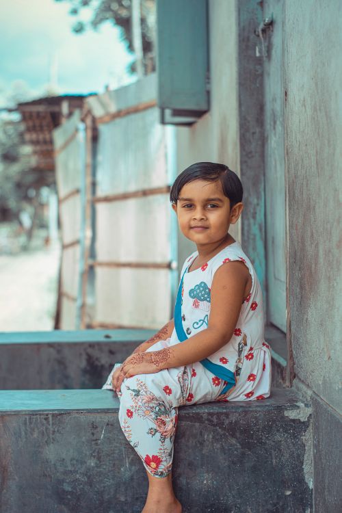 Free A smiling child with henna tattoo, wearing a floral dress, posing outdoors in a village scene. Stock Photo