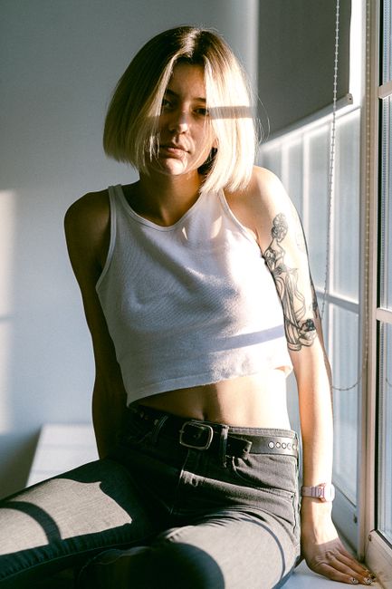 A tattooed woman wearing a white tank top sits by a sunlit window, showcasing her relaxed casual style.