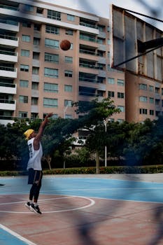A man wearing a yellow cap shoots a basketball on an outdoor court with urban buildings in the background.