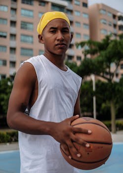 Portrait of a young man holding a basketball on an outdoor court in a city setting.