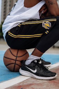 Man in sports attire sitting on a basketball on an outdoor court, close-up shot.