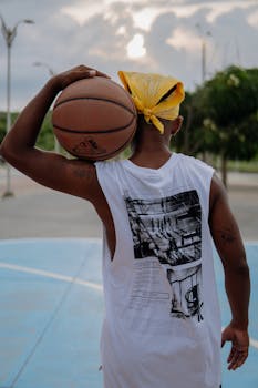 Basketball player holding ball on shoulder, standing confidently outdoors.