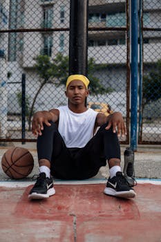 Young athlete resting on an outdoor basketball court next to a ball, wearing sports attire.