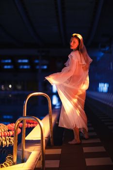A woman in a sheer dress stands by an indoor pool at night, illuminated by soft lighting.