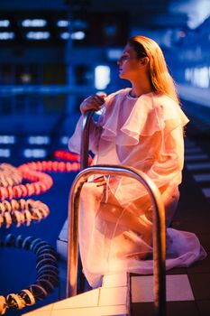 Stylish woman in a white dress kneeling by a pool embracing tranquility.