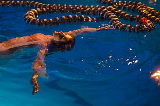 A woman floats calmly in a swimming pool under evening lights, surrounded by lane dividers.