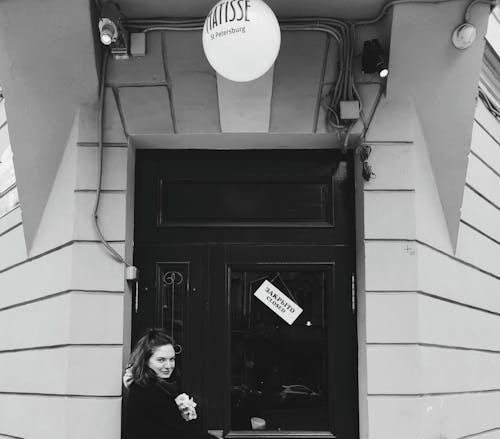 Grayscale Photo of Woman in Black Jacket Standing by a Door