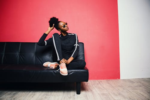 Femme Avec Pull Noir Et Blanc Avec Pantalon Assis Sur Un Canapé En Cuir Noir à Côté Du Mur Peint En Rouge