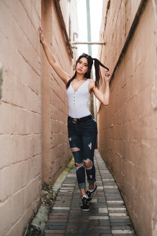 Woman in White Tank Top and Ripped Jeans Standing Between Concrete Walls