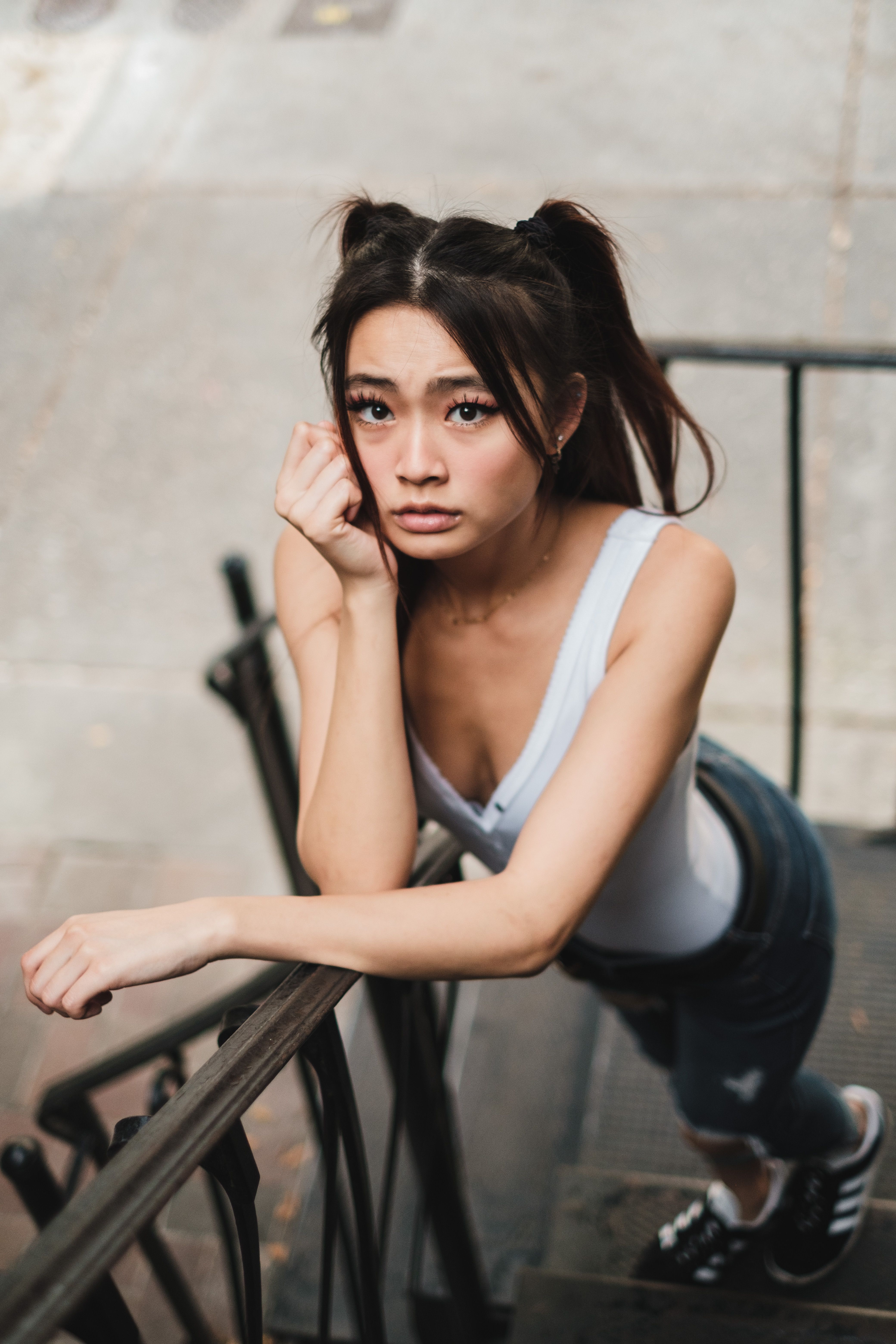 Asian woman in a white tank top leaning on a staircase outdoors, looking up.