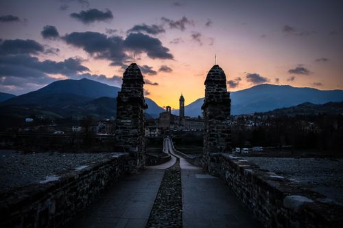 Free A picturesque medieval bridge leading to a historical town at sunset, surrounded by mountains. Stock Photo