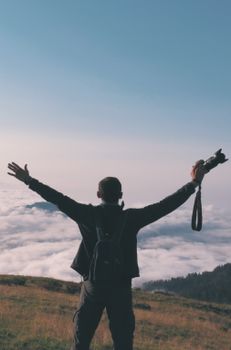 A man with arms outstretched, holding a camera, stands atop a hill above clouds during twilight, embracing freedom.
