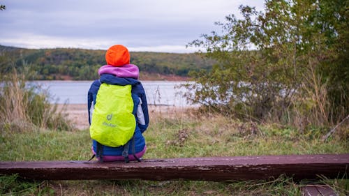 A Person in Blue Jacket with yellow Backpack Sitting Near a Lake