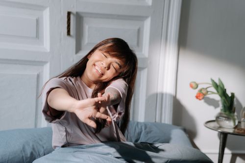 Free Smiling Asian woman stretching in bed, enjoying soft morning light indoors. Stock Photo