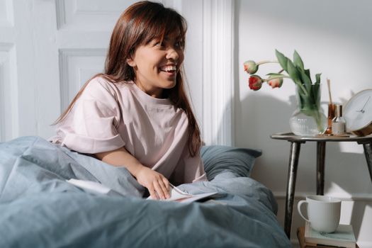 Asian woman with brown hair smiling, enjoying a calm morning in bed, surrounded by flowers and coffee.