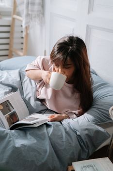 Asian woman relaxing in bed with a book and coffee. Indoors, cozy morning atmosphere.