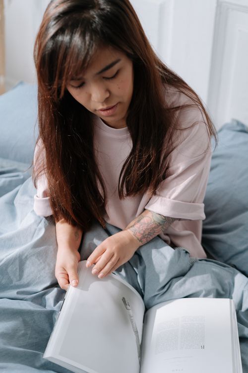 Free Asian woman reading a book in a cozy bedroom setting, enjoying a peaceful morning. Stock Photo