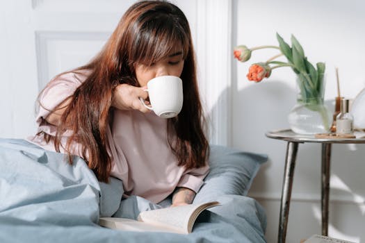 An Asian woman enjoying her morning with coffee and a book in a cozy bedroom setting.