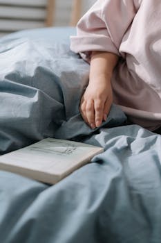 Close-up of a hand on bedding near an open book, conveying relaxation and quiet reading time.