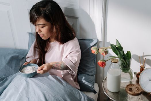 A woman sits in bed enjoying breakfast with milk and flowers nearby, bathed in morning light.