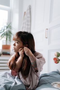 Woman with straight hair adjusting earrings in a serene bedroom setting.