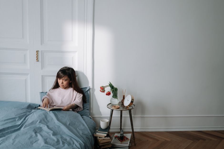 Woman sitting on a bed reading a book in a serene bedroom with morning light.