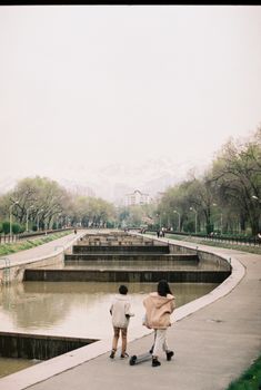 Children strolling along a scenic pathway in Almaty, with water features and trees.