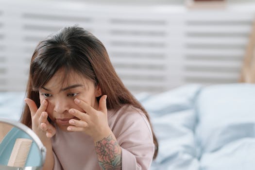 Asian woman applying skincare product in a bedroom setting with mirror reflection.