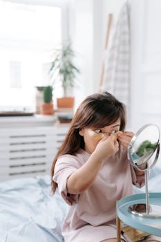 A young Asian woman applying eye patches in a bright, cozy bedroom setting.