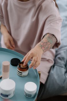 A woman with a tattooed arm reaches for face cream in a relaxing indoor setting.