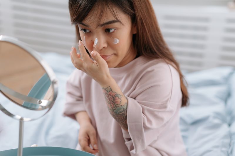 Adult woman applying face cream at home, embracing morning skincare routine in bedroom.