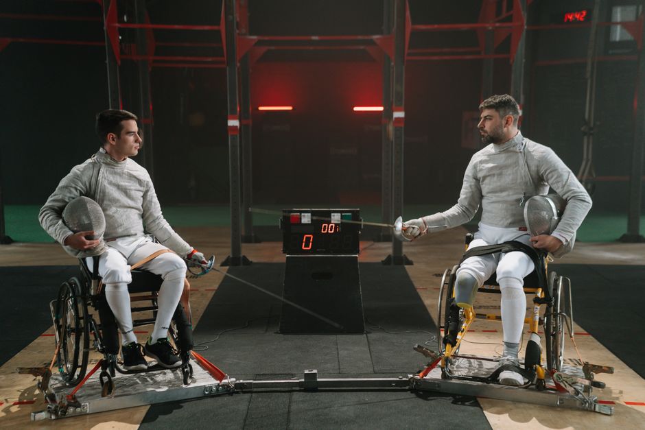 Adaptive fencing match between two athletes in wheelchairs, showcasing determination and skill indoors.